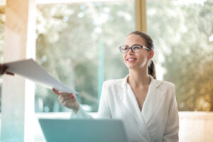 A woman in a white blazer receives a document from a person outside the frame, inside a bright office with a blurred background.