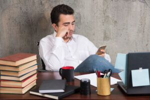 A person sits at a desk with books, a laptop, a mug, and stationery, engaged in using a smartphone in a stylish office setting.