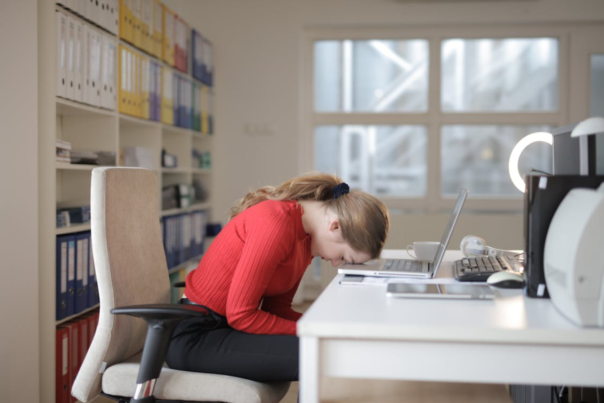 A young woman in a red sweater rests her head on a desk cluttered with a laptop.