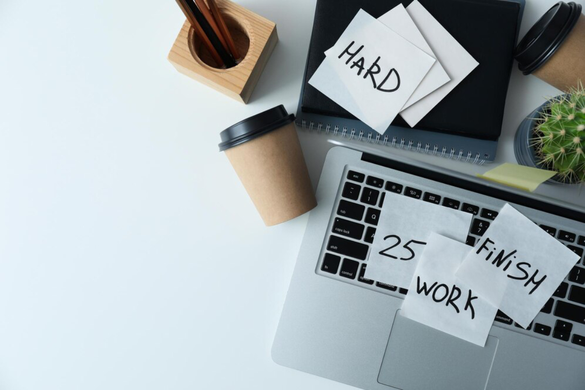 A laptop with sticky notes reading HARD, 25, FINISH, WORK, alongside a coffee cup, wooden pencil holder, and a cactus plant.