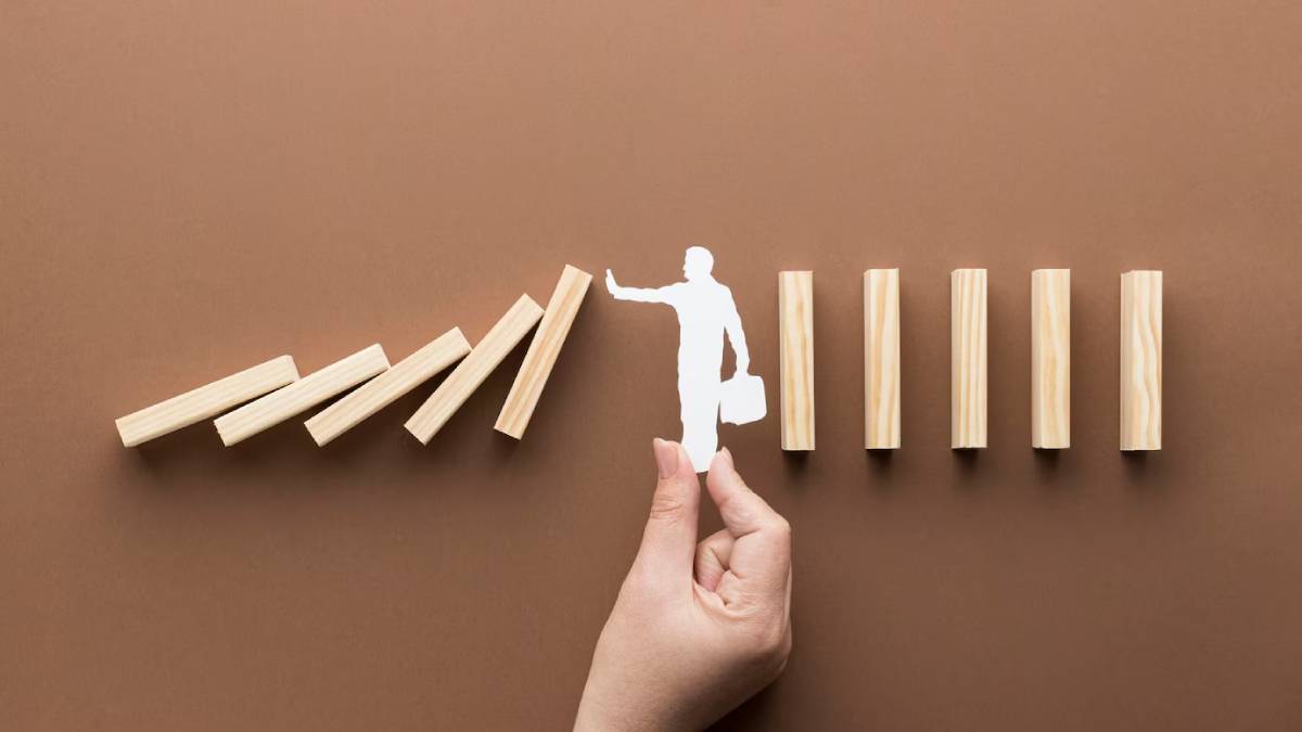 A hand holds a paper cutout of a person stopping a domino effect, on a brown background.