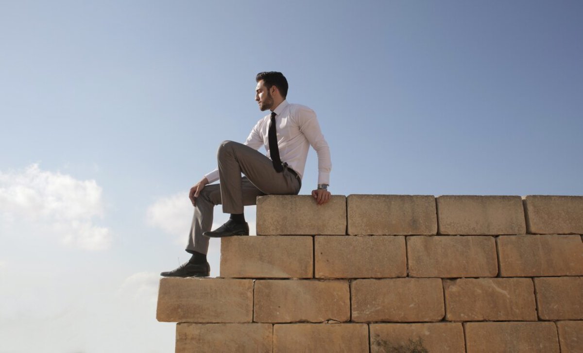 Man in business attire sits on a stone wall under a clear sky.