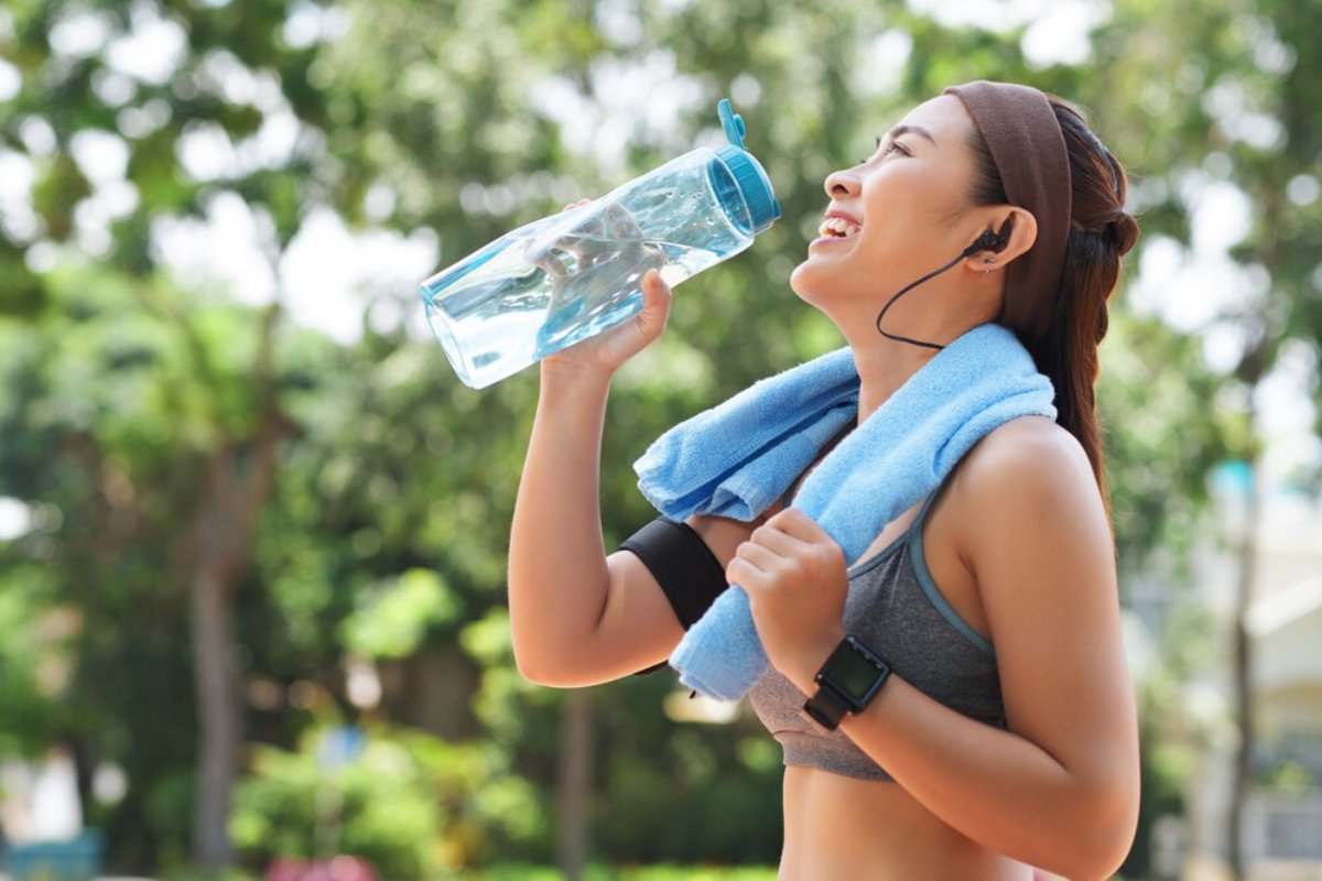 Woman in workout gear drinking water from a bottle outdoors, smiling with headphones and a towel around her neck.