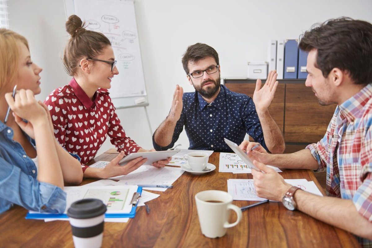 Four colleagues discussing graphs and documents at a conference table, one man gesturing emphatically.