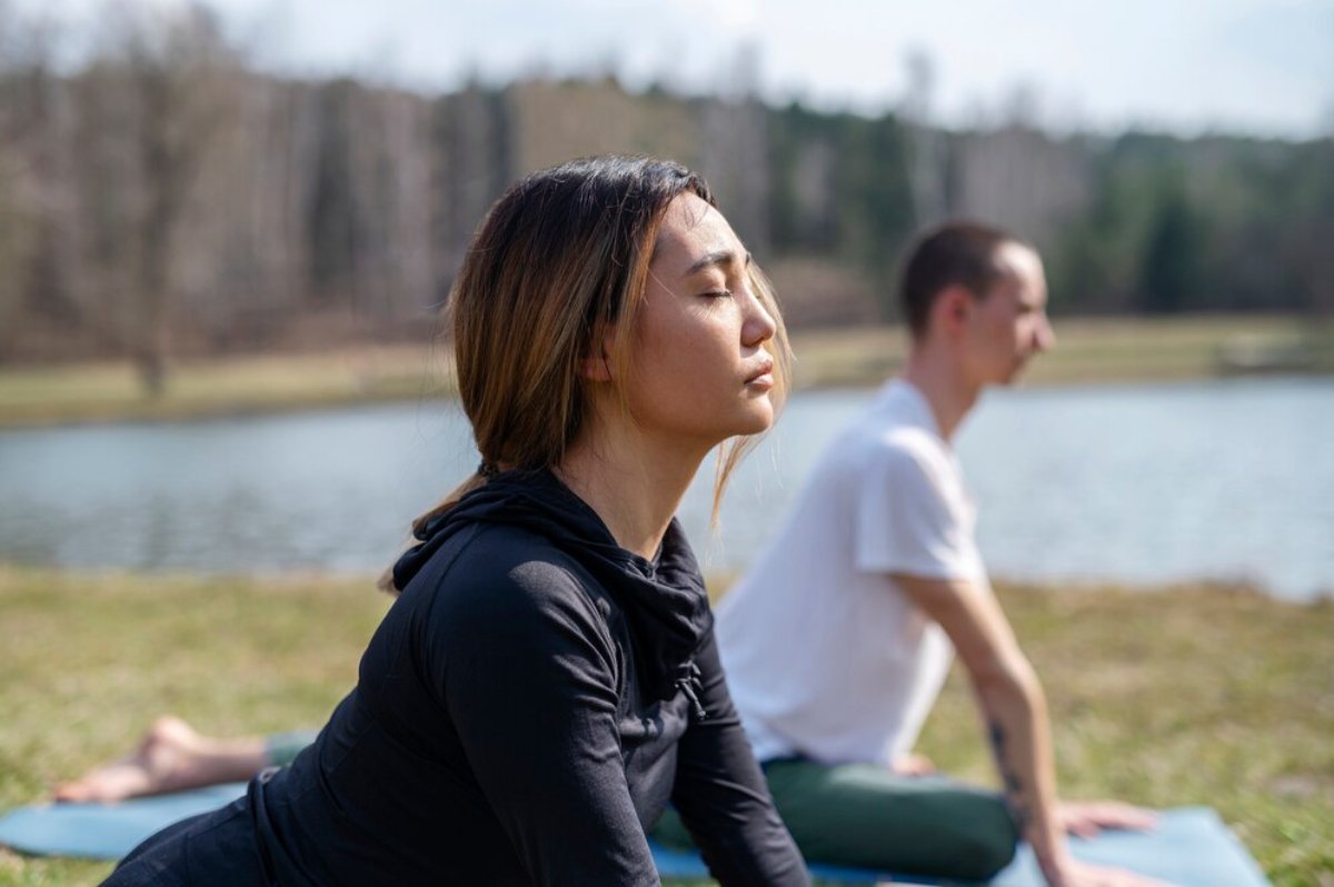 Two people practicing yoga outdoors near a lake, focusing peacefully with eyes closed.