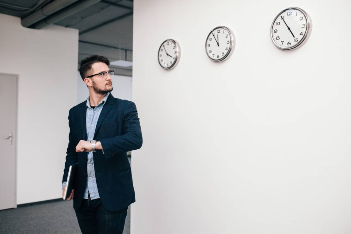 Man in business attire checking time on his wristwatch, standing beside a wall with three clocks.
