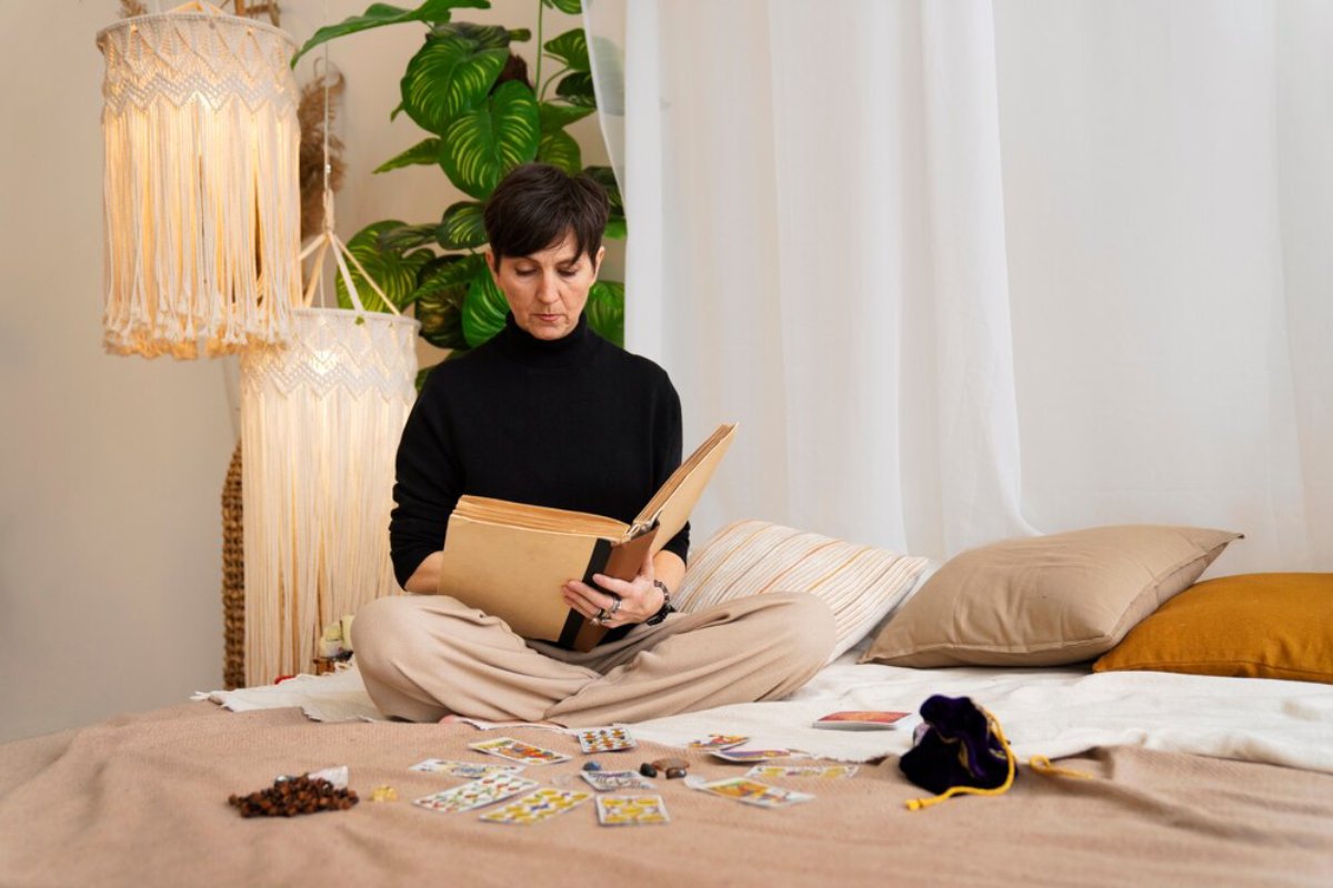 Woman reading a book while sitting on a bed with tarot cards and beads nearby, in a room with plants and a hanging lamp.