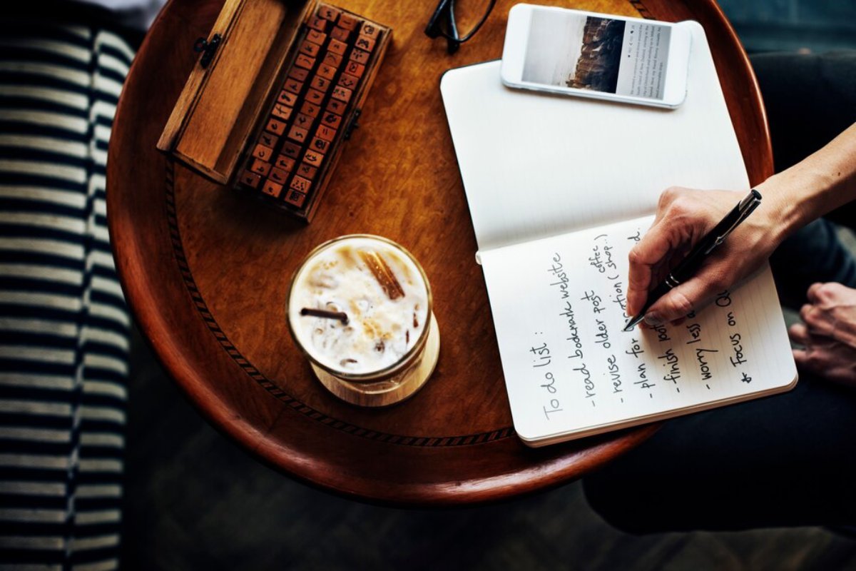Person writing a to-do list in a notebook on a wooden table with a smartphone and iced coffee nearby.