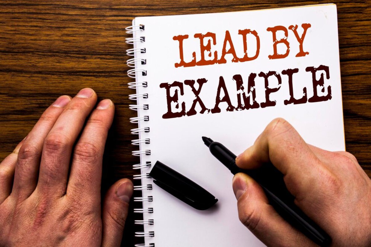 Person writing LEAD BY EXAMPLE in a notebook, with a black pen on a wooden desk.