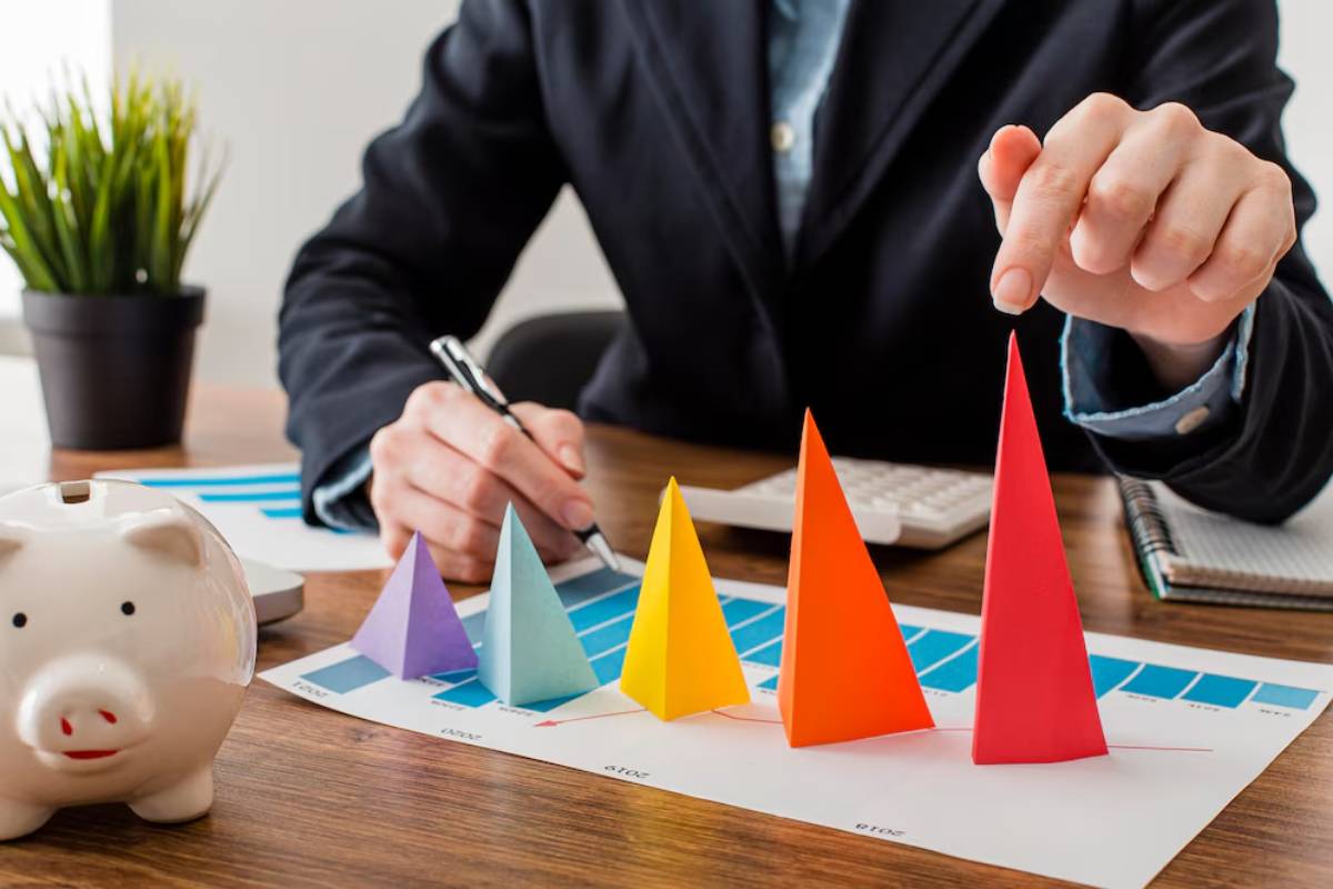 Man at a desk analyzes colorful geometric paper figures, with a piggy bank and financial charts surrounding him.