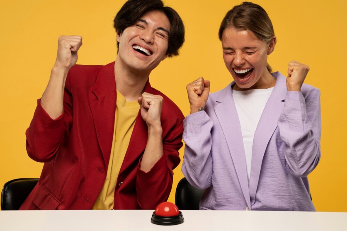 Two joyful people in colorful outfits celebrating a win, standing behind a table with a red buzzer on it, on a yellow background.