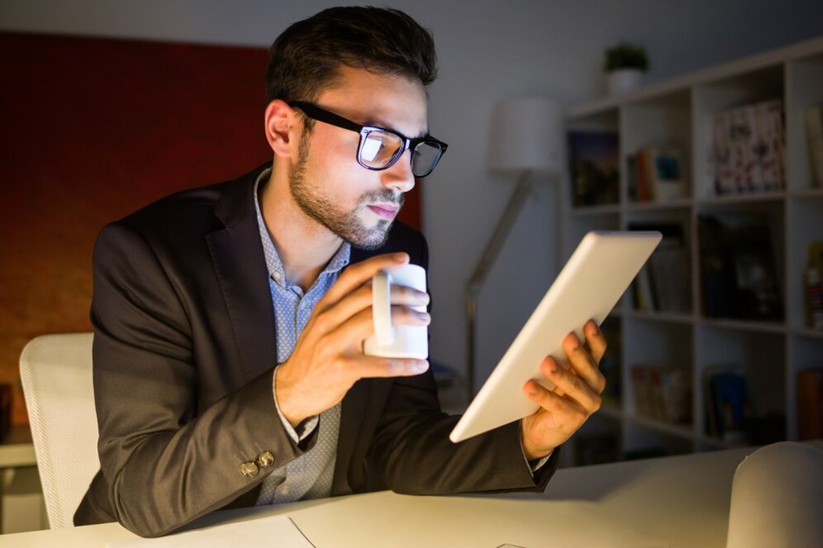 Young man in glasses using a digital tablet and drinking from a mug at a home office desk at night.