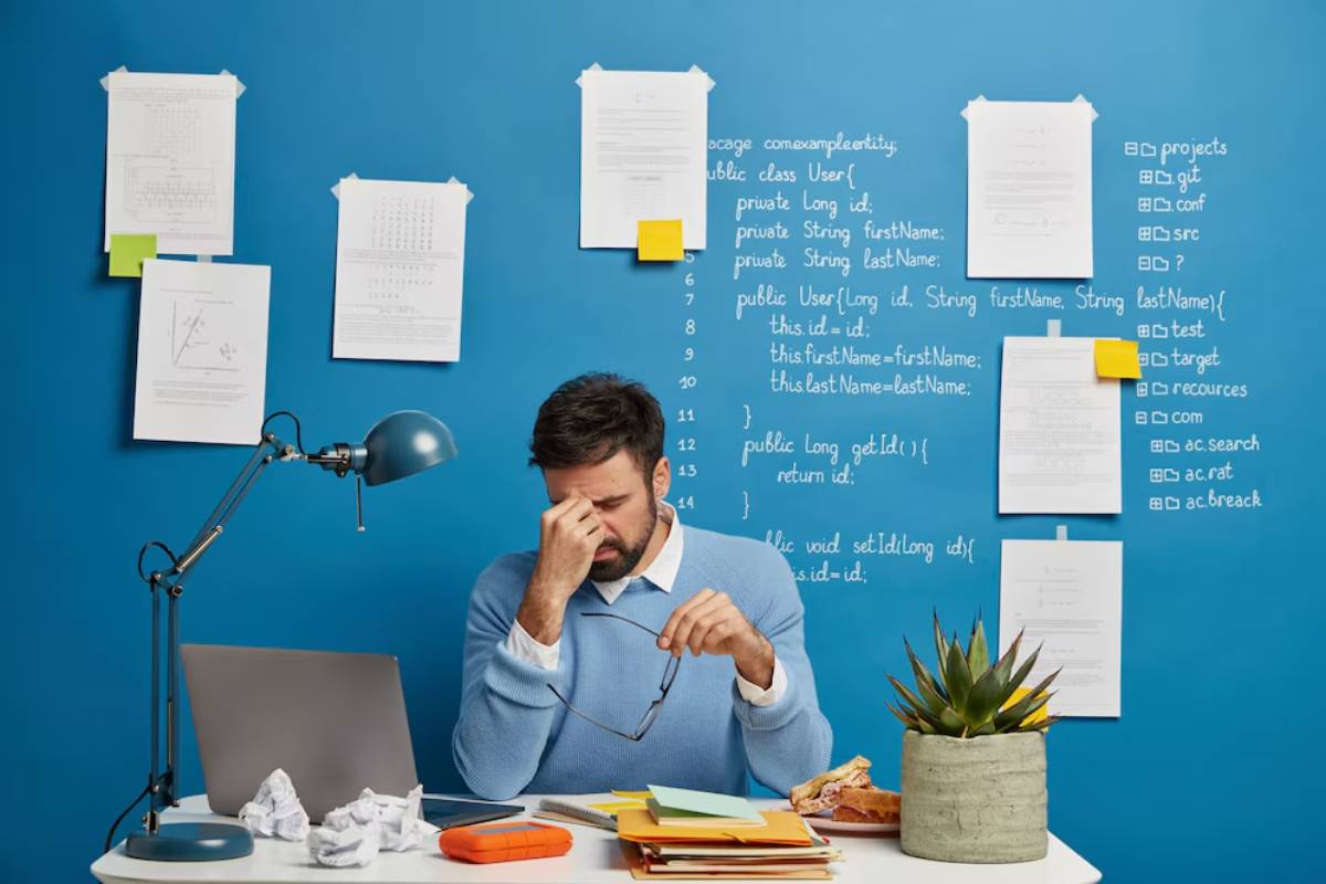 Man sitting at desk, looking stressed with paperwork and computer, blue wall covered with notes and diagrams in background.