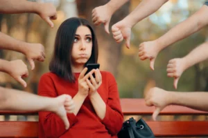 A woman in a red sweater sits on a bench, focused on her phone, surrounded by several hands showing thumbs down.
