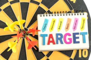 Dartboard with darts and a notebook labeled "TARGET" with colorful tabs reading goals like "Timely" and "Achievable."