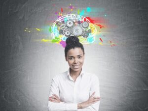 A woman with arms crossed, smiling in front of a chalkboard with a colorful, mechanical brain illustration above her head.