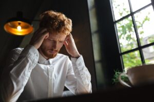 A distressed young man with red hair sitting indoors, holding his head in his hands.