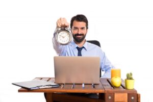 Businessman at desk holding a clock, smiling, with a laptop, fruit, and juice on his desk.