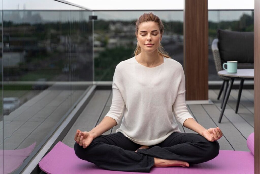 Woman meditating on a balcony, wearing a white top and black pants, calm cityscape in the background.